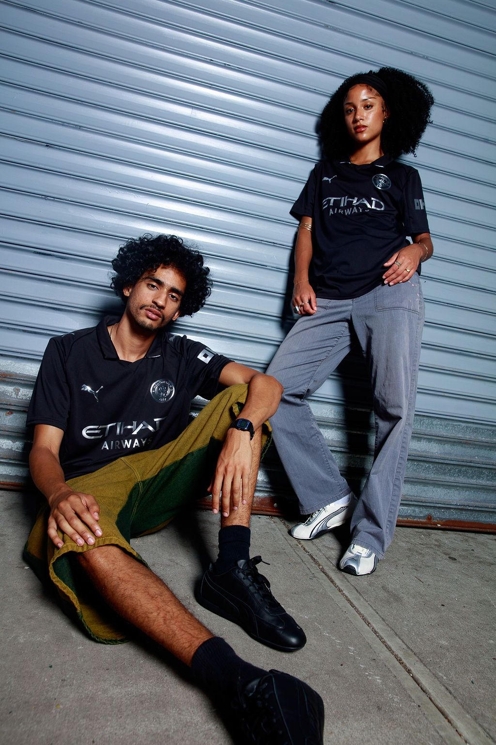 Two models in black Manchester City shirts beside a shuttered storefront