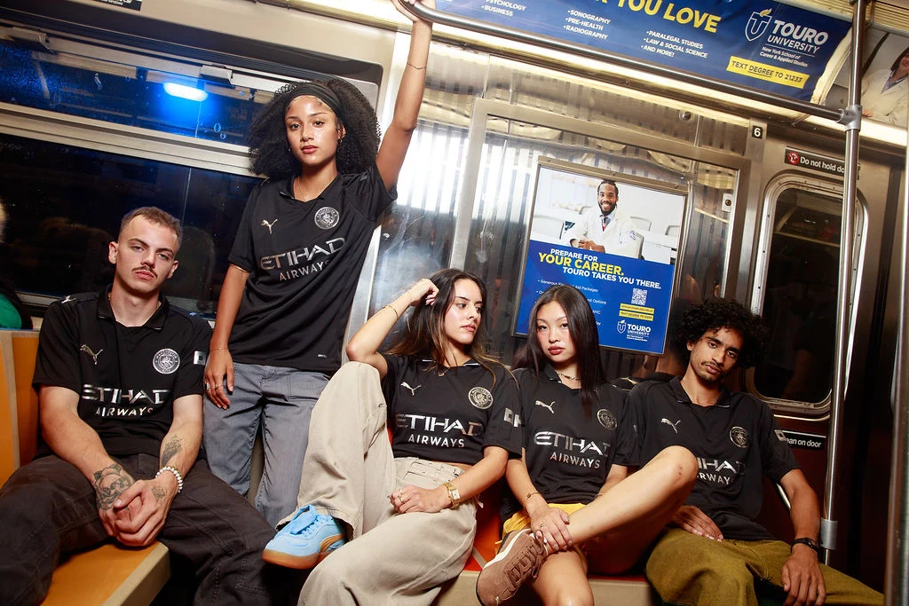 Five models wearing black Manchester City away kits inside a New York City subway car